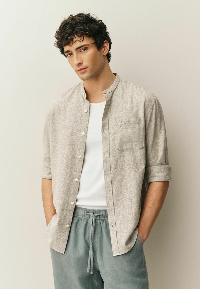 Camicia grigio chiaro a maniche corte con bottoni, colletto e tasca sul davanti, indossata sopra una canottiera bianca. Abbinata a pantaloni larghi di colore azzurro chiaro.