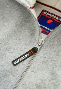 Felpa grigia con zip, caratterizzata da una cerniera metallica, etichetta nera con il marchio "SUPERDRY" e un rivestimento interno morbido con un'etichetta che mostra grafiche colorate.