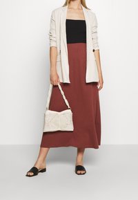 Blazer beige, top sans manches noir, jupe maxi couleur rouille, sac à main tissé blanc et sandales à enfiler noires. Tissu texturé et détails de bretelles tressées.