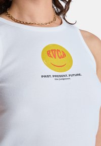 Débardeur blanc côtelé avec un motif de smiley jaune, accompagné du texte rouge "RVCA" et "PASSÉ. PRÉSENT. FUTUR." imprimé en dessous. Collier chaîne dorée.