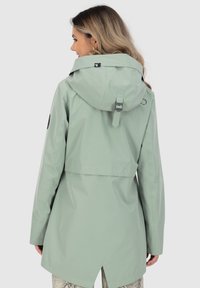 Hellgrüne wasserdichte Jacke mit Kapuze, verstellbarem Riemen und Belüftung. Mit einem seitlichen Schlitz auf dem Rücken und einem runden schwarzen Logo-Element am Ärmel.