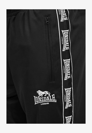 Czarne sportowe spodnie z białym logo Lonsdale na lewej uda. Posiadają kieszeń na zamek oraz czarno-białe paski po bokach.