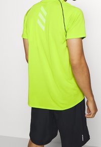 Neon gul sporttröja med en ventilerande mesh-textur, försedd med tre reflexstreck på ryggen, tillsammans med svarta träningsshorts.