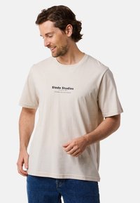 Beige kortærmet t-shirt med rund hals, der har et sort logo "Slade Studios" på forsiden. Lavet af bomuldsstof med en glat tekstur.