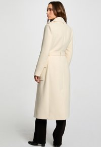 Crème-kleurige wollen jas met een ceintuur om de taille, zijzakken en een hoge kraag. Heeft een getailleerde pasvorm en een middellange lengte.