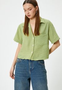 Camisa verde claro de manga corta con un acabado texturizado, con botones en la parte delantera y un bolsillo en el pecho, combinada con unos pantalones vaqueros azules de cintura alta.