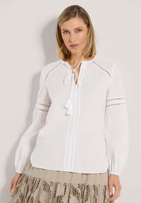 Blonde Frau, die eine weiße Bluse mit langen Ärmeln und Spitzenmotiven sowie einer Quastenbindefunktion trägt, kombiniert mit einem beigen Rock mit gestickten Mustern.