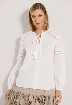 Blonde Frau, die eine weiße Bluse mit langen Ärmeln und Spitzenmotiven sowie einer Quastenbindefunktion trägt, kombiniert mit einem beigen Rock mit gestickten Mustern.