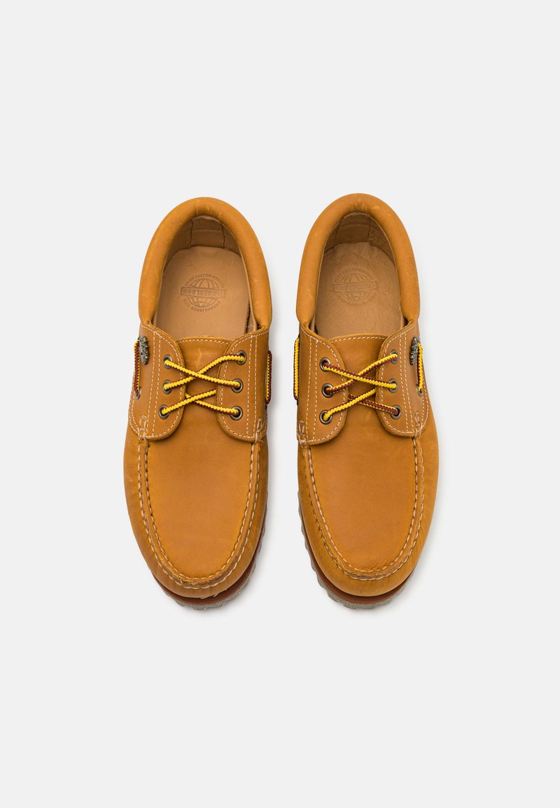 Een paar tan leren bootschoenen met gele veters, gestikte details en rubberen zolen, van bovenaf bekeken op een witte achtergrond.