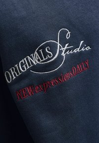 Σκούρο μπλε ύφασμα με λευκή κεντημένη επιγραφή "ORIGINALS Studio" και κόκκινη κεντημένη επιγραφή "NEW expressions DAILY" σε κυρτή επιφάνεια.