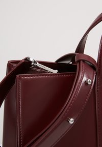 Bordeauxrode leren handtas met een gestructureerd ontwerp, voorzien van zilverkleurige hardware en een ritssluiting. Gladde textuur met zichtbare stiksel details.