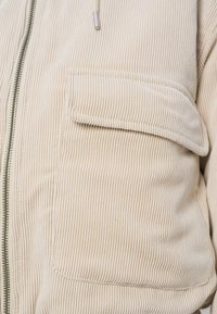 Μπεζ βαμβακερό μπουφάν με φερμουάρ, με κάθετη ριγωτή υφή, μπροστινή τσέπη και κουκούλα με κορδόνια. Διακοσμητικά στοιχεία περιλαμβάνουν ασημένιο φερμουάρ.