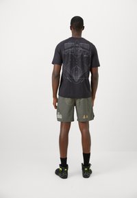 Camiseta gráfica negra con un diseño blanco en la parte posterior, combinada con pantalones cortos verdes oliva que presentan varios patrones y acentos dorados. Calzado negro.