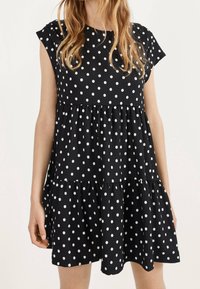 Kvinna klädd i en svart, ärmlös, lager-på-lager klänning med vita prickar, stående mot en ljus, enfärgad bakgrund.