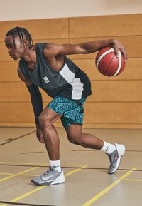 Joueur de basket dribblant un ballon, portant un t-shirt sans manches gris, un short imprimé léopard teal, des chaussures grises avec des accents roses et des chaussettes blanches.