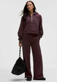 Bordo zip-up skrajšan hoodie z sprednjim žepom, v kombinaciji z ohlapnimi bordo hlačami. Drži črn nahrbtnik z vrvico; raven črni čevlji.