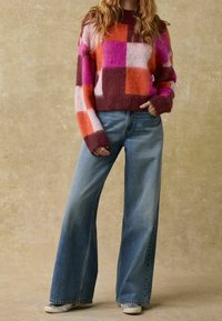 Bunt karierter Pullover in Rosatönen, Orange und Braun, kombiniert mit weit geschnittenen hellen Jeans und grauen Sneakers.