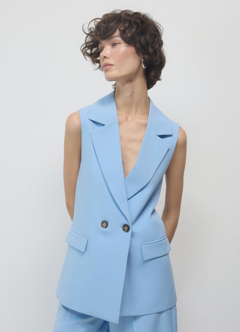 Blazer senza maniche blu chiaro con profondo scollo a V, chiusura a doppio bottone e due tasche frontali. Texture liscia e design strutturato.