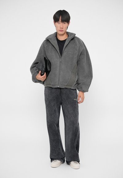Graue, flauschige Zip-Jacke mit strukturierter Oberfläche, kombiniert mit dunkelgrauen Fleece-Hosen und weißen Sneakers. Schwarze Clutch in der Hand.