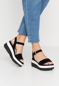 Sandali platform in suede nero con una suola intermedia bianca, cinturino alla caviglia regolabile, design a punta aperta e suola in gomma texturizzata.