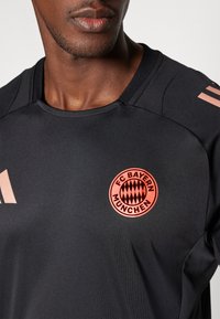Sort atletisk skjorte med rund halsudskæring, der har et cirkulært FC Bayern München-logo i pink med et diamantmønster og pink striber på skulderen.
