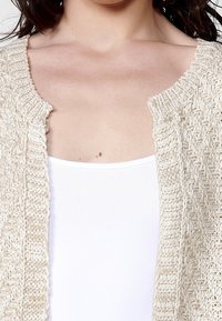 Beżowy dziergany kardigan o fakturowanym splocie, zapinany z przodu na otwartą część, o miękkim i lekkim wyglądzie. W zestawie z białym, bezrękawnym topem pod spodem.