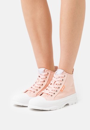 Jalat pukeutuneina vaaleanpunaisiin korkeavartisiin kangaskenkkiin, joissa on valkoiset kumipohjat ja nauhat, seisomassa yksinkertaisella vaalealla taustalla.