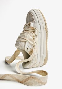 Beige sneaker met een gestructureerde stof bovenwerk, gewatteerde kraag, platte veters en een geprinte veter. Dikke zool met een lichte kromming in vorm.
