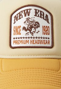 Gorra beige con parche bordado que presenta "NEW ERA", un jinete sobre un caballo y "SINCE 1920" en marrón, con visera amarilla texturizada.