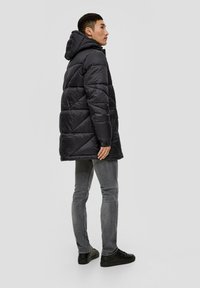 Chaqueta acolchada negra con capucha, con un patrón de acolchado, bolsillos laterales y un corte relajado. Combinada con jeans grises y zapatos negros.