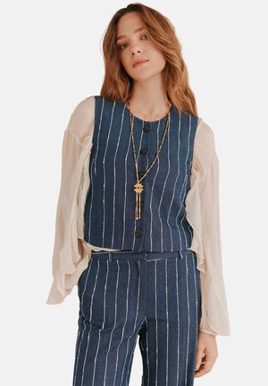 Denimvest med lodrette hvide striber, fem knapper, parret med lette, gennemsigtige lange ærmer. Guldkæde med blomst vedhæng.