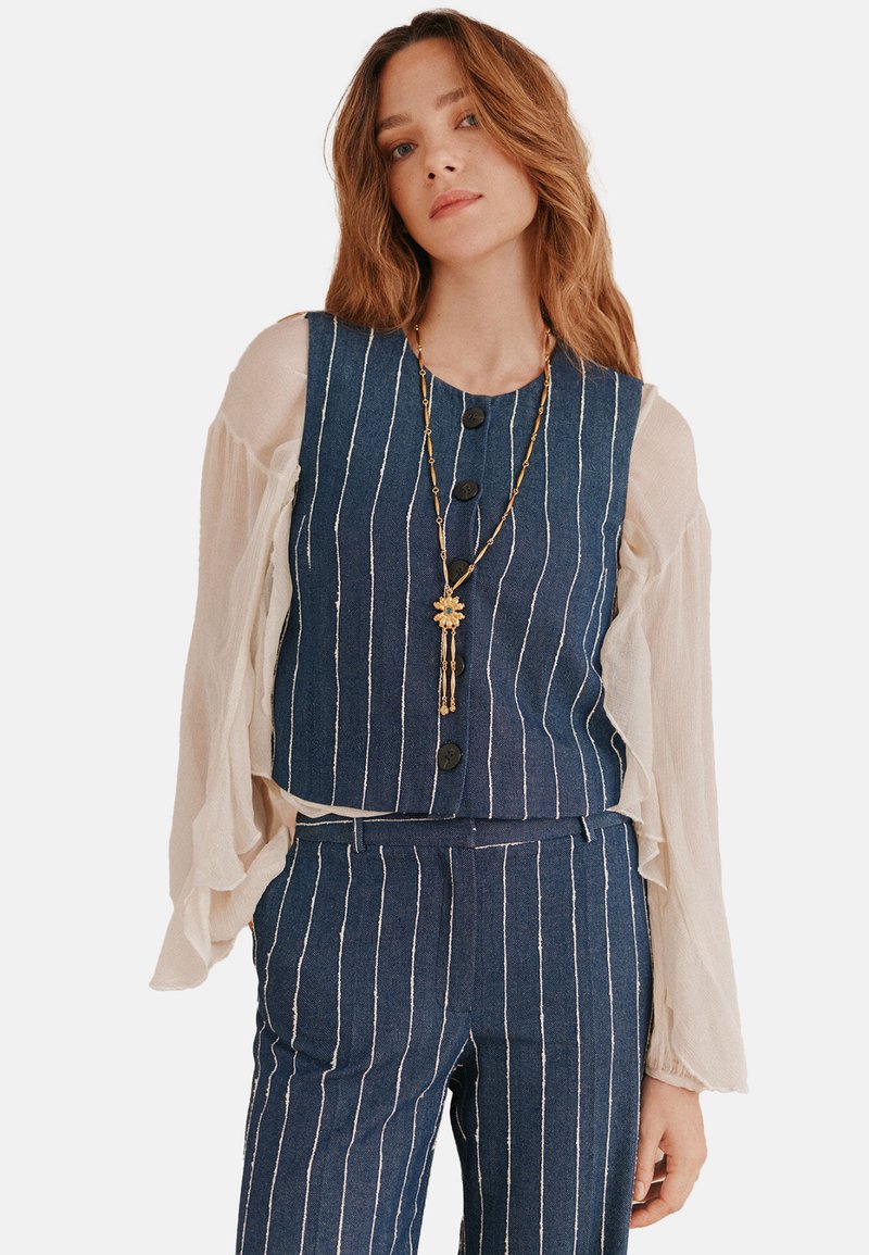 Gilet di jeans con righe bianche verticali, cinque bottoni, abbinato a maniche lunghe fluenti e trasparenti. Collana in oro con pendente a forma di fiore.