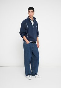 Sweatshirt de lã polar azul-marinho com fecho de um quarto, detalhes brancos e colarinho alto, combinado com calças de ganga largas azuis e ténis brancos.