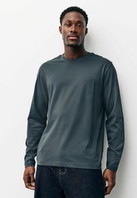 Mies lyhyillä kiharoilla hiuksilla, yllään tummanharmaa pitkähiha-paita ja tummat farkut, seisoo valkoista taustaa vasten katsellen eteenpäin.