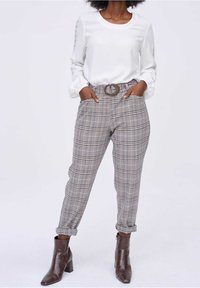 Femme portant un chemisier blanc à manches longues, un pantalon à carreaux taille haute avec ceinture, et des poignets retroussés, associé à des bottines marron.