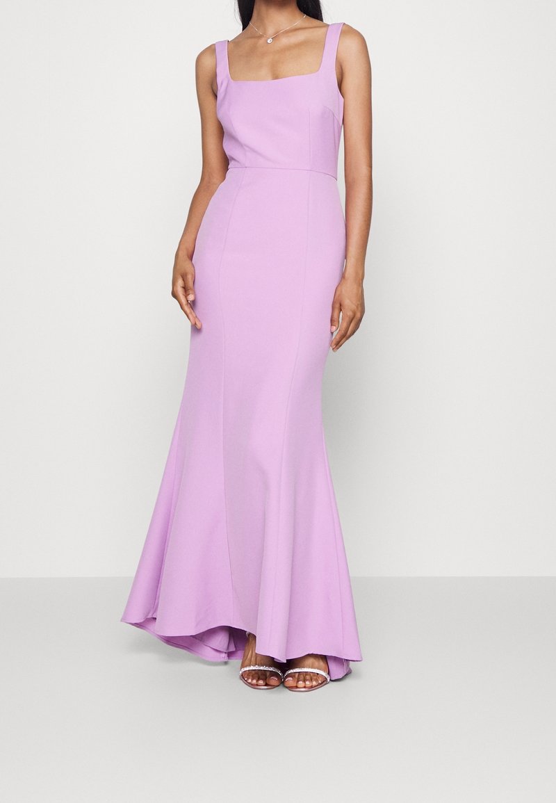 Femme portant une robe de soirée lilas ajustée sans manches avec un décolleté carré et un ourlet évasé, associée à des talons ouverts argentés.