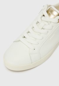 Sneaker in pelle bianca con punta tonda, suola piatta in gomma e chiusura con lacci. Presenta un accento con logo dorato e un pannello laterale texture.