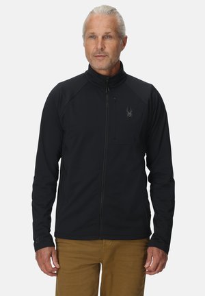 Mann mit grauen Haaren, der eine schwarze Jacke mit Reißverschluss und einem Spinnenlogo auf der Brust sowie braune Hosen trägt, vor einem weißen Hintergrund stehend.