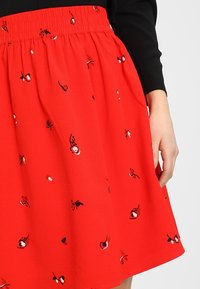 Jupe rouge avec taille élastique, décorée de broderies florales éparpillées en blanc et bleu marine. Tissu léger, forme évasée, longueur au genou.