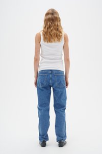 Weißes geripptes Tanktop, blaue gerade Jeans aus Denim, sichtbare Nähte und Gesäßtaschen. Person, die sich abwendet, trägt schwarze Schuhe.