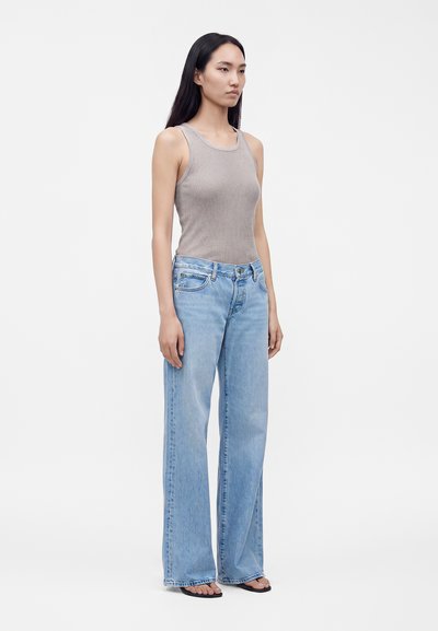 Femme debout portant un top gris sans manches côtelé, un jean large bleu clair et des sandales noires sur un fond clair uni.