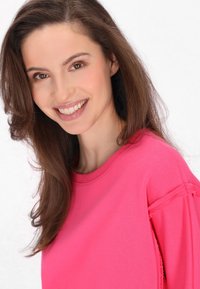 Felpa di un vivace colore rosa con scollo a girocollo, tessuto morbido e dettagli sfrangiati sulle maniche. Modella sorridente su sfondo bianco.