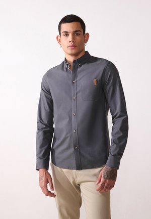 Camisa gris de manga larga con botones y cuello, que presenta un bolsillo en el pecho y botones marrones. Textura suave y diseño ajustado.