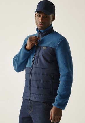 Mężczyzna w niebieskim polarze i pikowanej kurtce, zapinający zamek błyskawiczny, w ciemnoniebieskiej czapce i dżinsach na jednolitym tle.