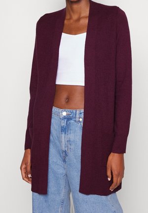 Cardigan violet foncé ouvert porté sur un débardeur blanc court et un jean bleu clair taille haute.