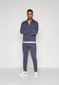 Homem de pé com os braços cruzados, vestindo um fato de treino azul marinho e ténis brancos, contra um fundo claro liso.