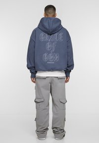 Marineblauwe oversized hoodie met grote witte letters op de achterkant, gecombineerd met lichtgrijze cargobroek met zijzakken en een losse pasvorm.
