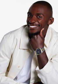 Round watch with a black dial, gold and silver metal band, displayed on a hand resting under a chin. Man wearing a cream jacket and white shirt.