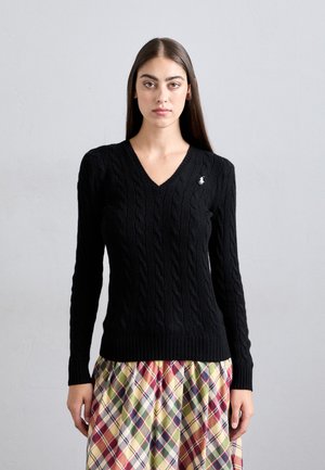 Woman with long dark hair wearing a black cable-knit V-neck sweater and a multicolored plaid skirt, standing against a plain light gray background.