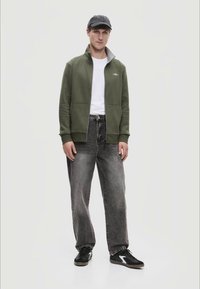 Olíva zöld, cipzáros pulóver, fehér póló, szürke, bő fazonú farmernadrág, fekete sportcipő fehér részletekkel és sötét sapka. Simább textúra és laza szabás.